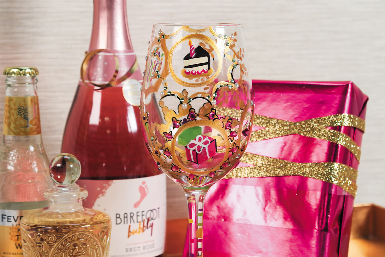 Decorative wine glass with festive design, pink bottle, and gift box on a neutral background