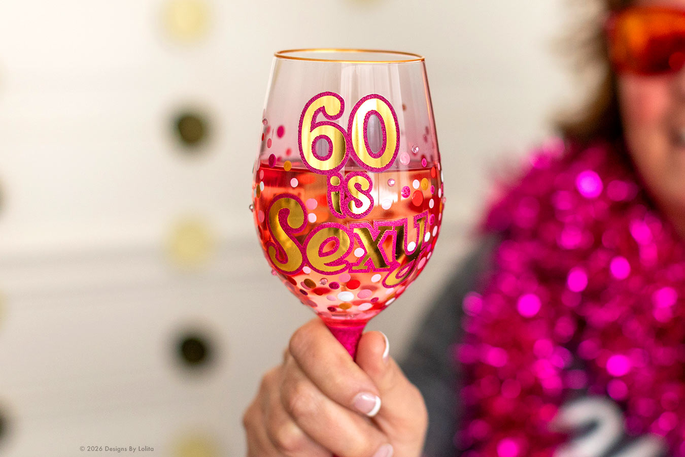 A hand holds a clear wine glass decorated with pink glitter, red polka dots, and the phrase '60 is Sexi' in yellow and pink lettering. A person wearing a bright pink tinsel garland is blurred in the background.