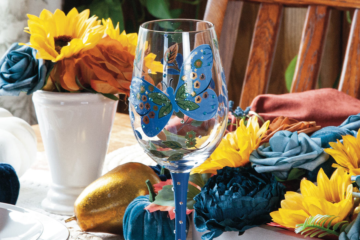 A clear wine glass with a hand-painted blue butterfly and blue stem, styled on a table with autumn flowers, a white vase, and a wooden chair in the background.